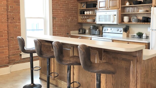 Una cocina con pared de ladrillo, armarios de madera, encimera blanca y cuatro taburetes de bar.