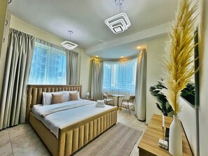 A modern hotel room with a large bed, two bedside tables, a small round table, and a view of the city through large windows. - Lux Marina-View 2 Bedroom Unit, Walking Distance to JBR Beach, Metro & Tram. (Dubai)