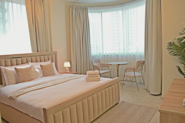 A hotel room with a large bed, a bedside table with a lamp, a small round table with chairs, and a potted plant. - Lux Marina-View 2 Bedroom Unit, Walking Distance to JBR Beach, Metro & Tram. (Dubai)