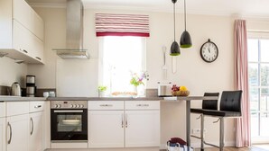 A modern kitchen with white cabinets, a built-in oven, a coffee maker, and a clock on the wall. - Ferienhaus Schilfrohrsanger, Fuhlendorf (Fuhlendorf)