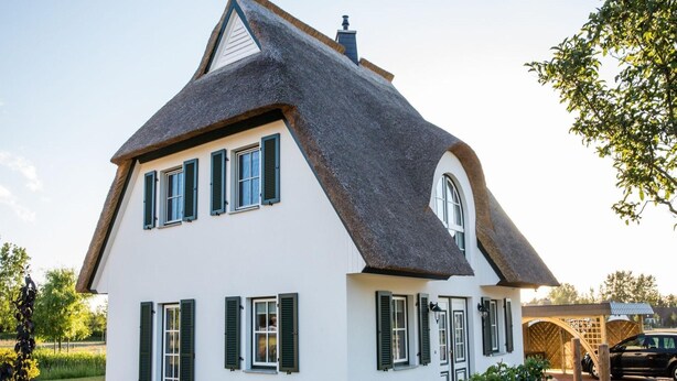 A two-story house with a steeply pitched roof, multiple windows with shutters, and a well-maintained garden with gravel pathways and greenery. - Ferienhaus Schilfrohrsanger, Fuhlendorf (Fuhlendorf)
