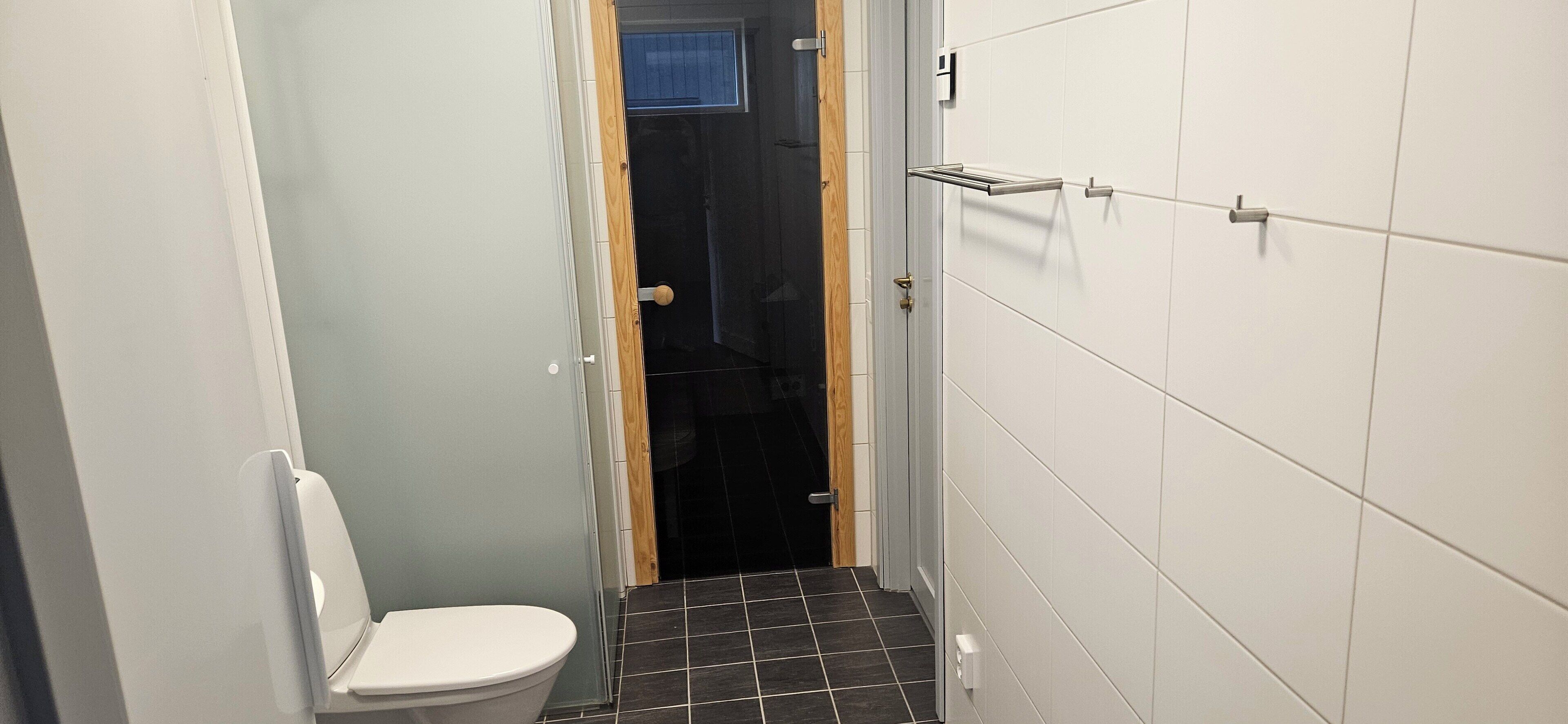 A narrow bathroom with a white toilet, black tiled floor, and white tiled walls.