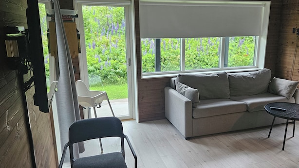 A cozy room with a wooden floor, a grey sofa, a small round table, and a window with a view of greenery. - Skidvägen 4a - Lodge (Jarvso)