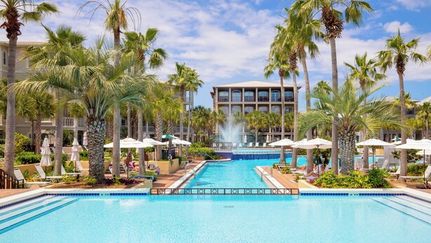 A swimming pool with lounge chairs and umbrellas, surrounded by palm trees and a building in the background.