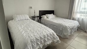 A hotel room with two single beds, a nightstand with a lamp, and a window with curtains. - Estudio Urbano en Santiago (Santiago)