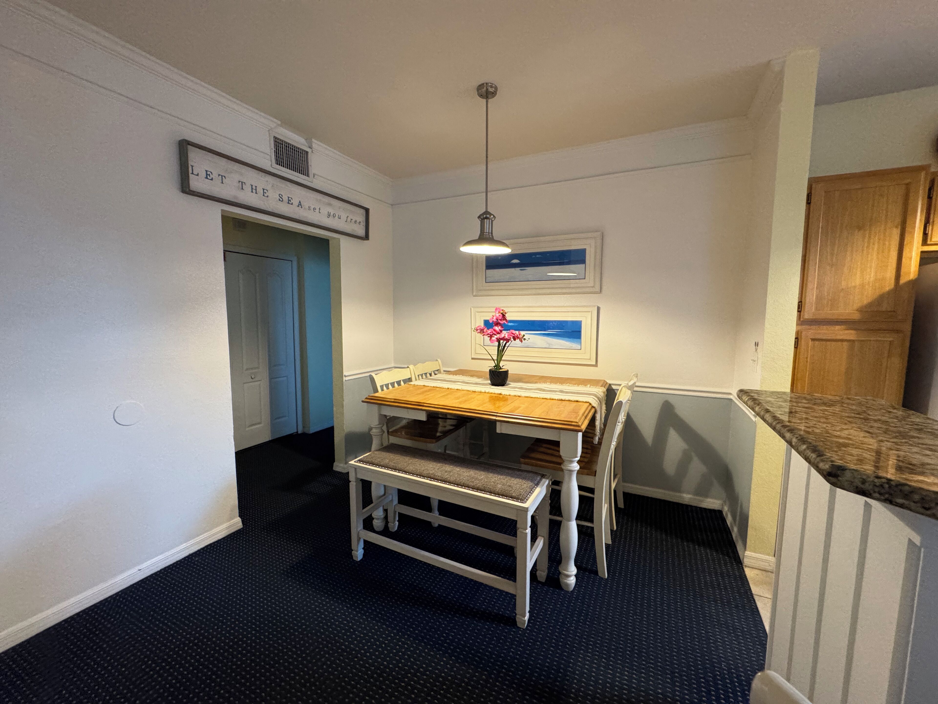 A small dining area with a wooden table and chairs, a hanging light fixture, and a framed picture on the wall.
