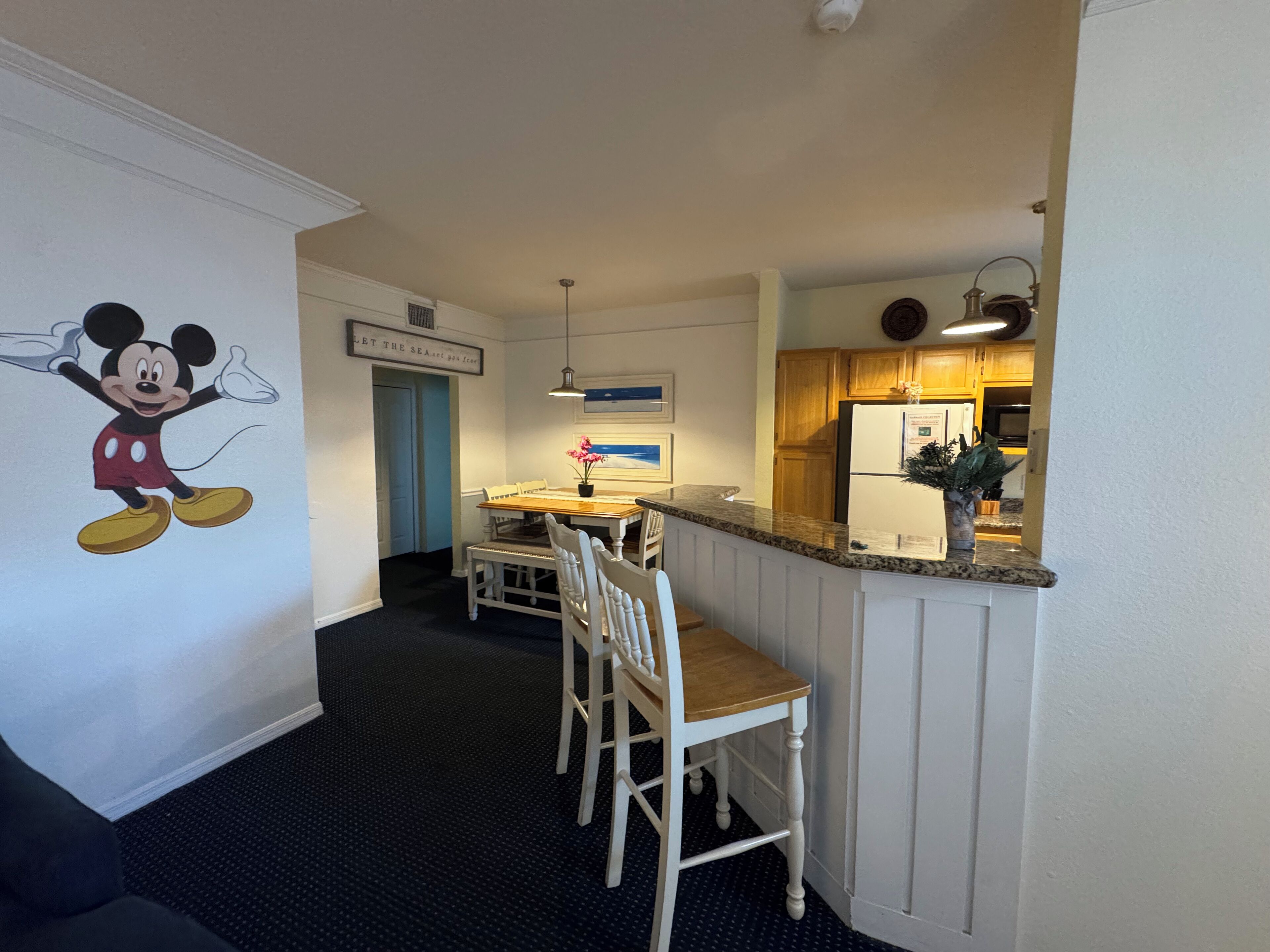 Habitación de hotel con un mural de Mickey Mouse, una zona de comedor con mesa y sillas, una cocina con armarios de madera y una puerta azul.