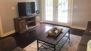 Een woonkamer met een donker houten vloer, een televisie op een houten meubel, een bijzettafeltje met decoratieve objecten, en een grijze bank.