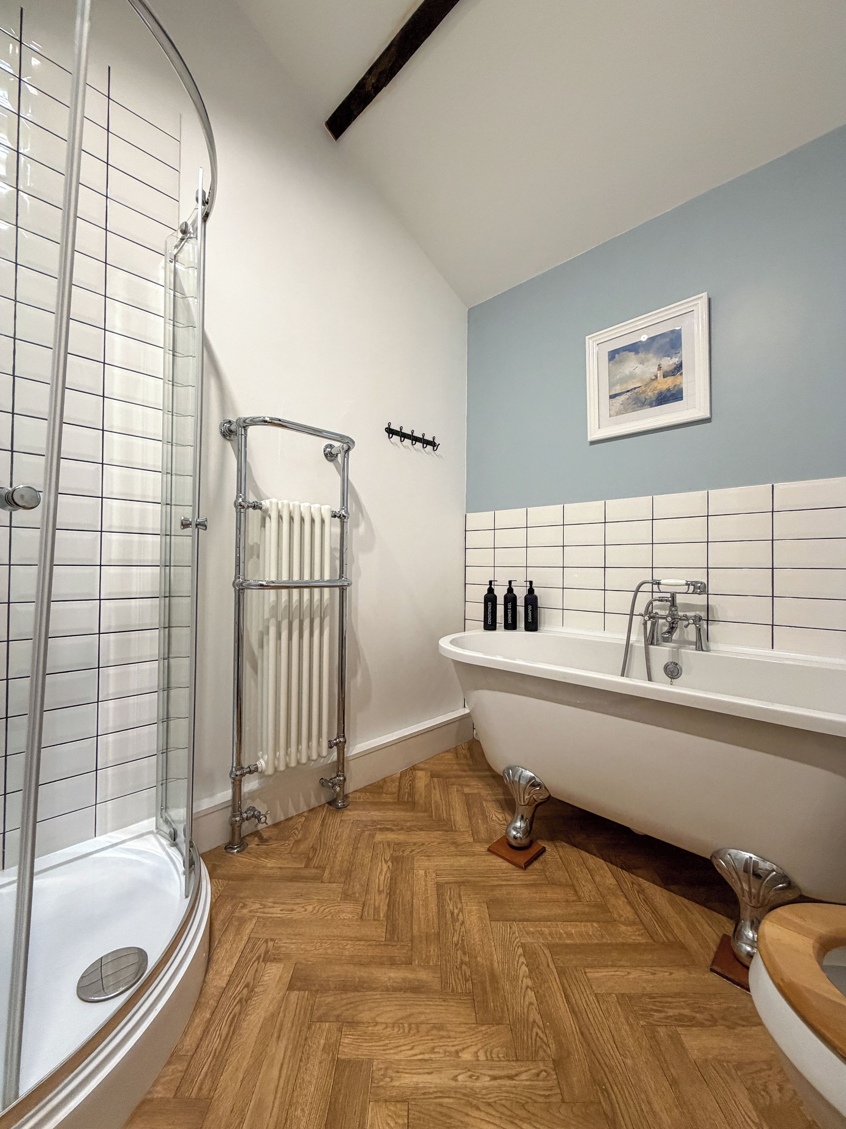 A bathroom with a glass shower enclosure, a freestanding bathtub, a white radiator, and a tiled wall with a painting.