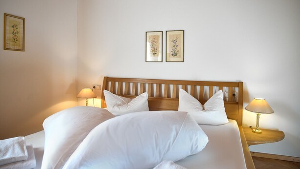 Una cama bien hecha con sábanas y almohadas blancas, dos lámparas fijadas a la pared y dos cuadros con motivos florales.