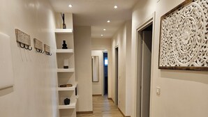 Un couloir doté d’un plancher en bois, de murs blancs et d’un panneau décoratif sur le côté droit. Des bancs blancs sont disposés le long du mur gauche, et une porte se trouve au fond.