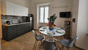 Une cuisine moderne avec des meubles de couleur sombre, un four encastré et un four à micro-ondes. On y trouve une table à manger avec quatre chaises et une plante en pot. Un téléviseur est fixé au mur, et une fenêtre laisse entrer la lumière naturelle.