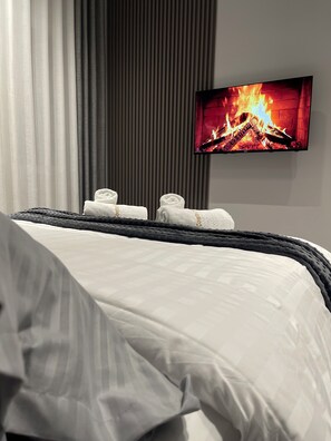 A hotel room with a bed, a flat-screen TV displaying a fireplace, and a window with curtains.
