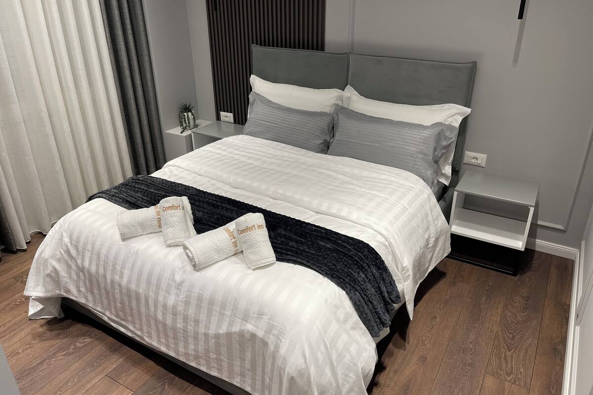 A neatly made bed with a striped duvet, pillows, and a dark throw blanket. There is a small bedside table with a lamp and a power outlet.