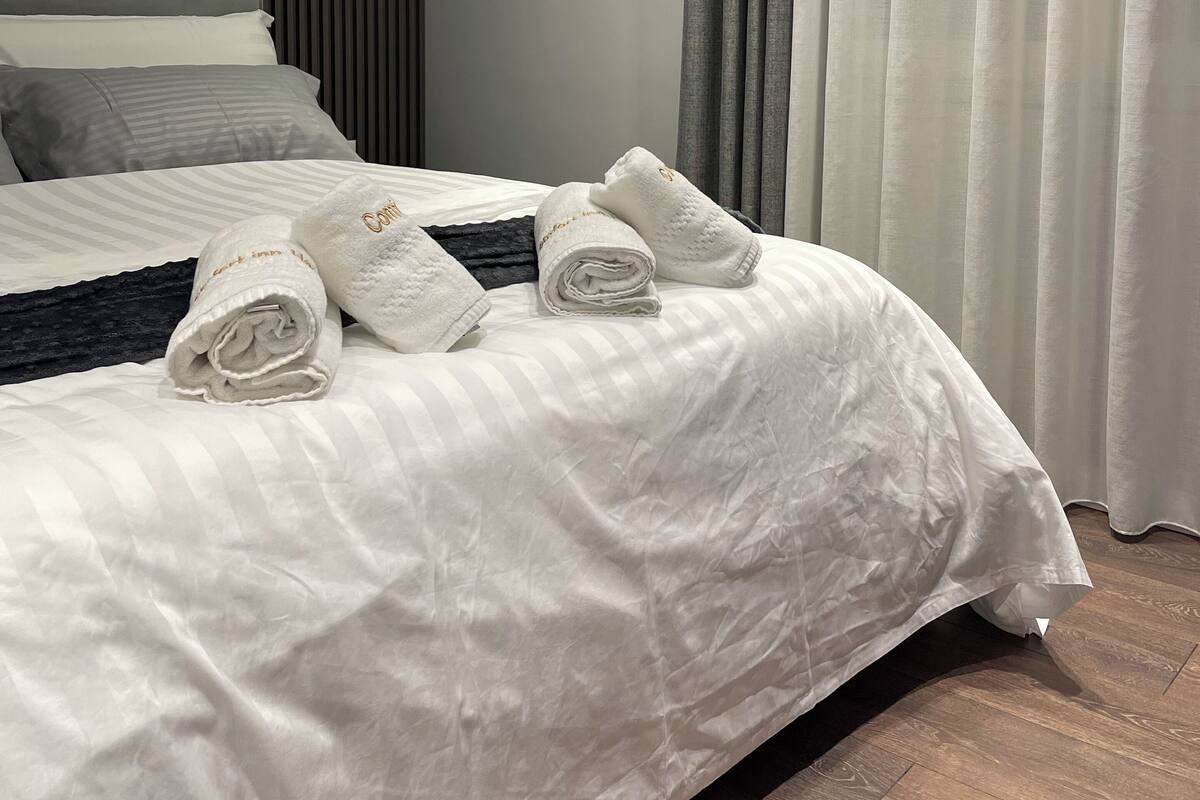 A neatly made bed with white linens and rolled towels, a grey headboard, and a grey curtain.