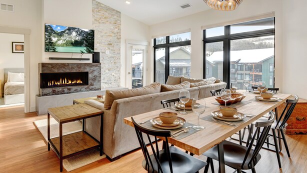 A modern living room with a fireplace, a dining table set for six, and a large window offering a view of a snowy landscape.