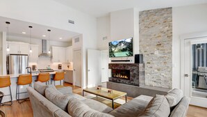 A modern living room with a large sofa, a coffee table, and a fireplace. There is a kitchen area with white cabinets and a stainless steel refrigerator. A flat-screen TV is mounted above the fireplace.