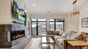 A modern living room with a fireplace, a large TV mounted on the wall, a sofa, a coffee table, and large windows offering a view of buildings and mountains.