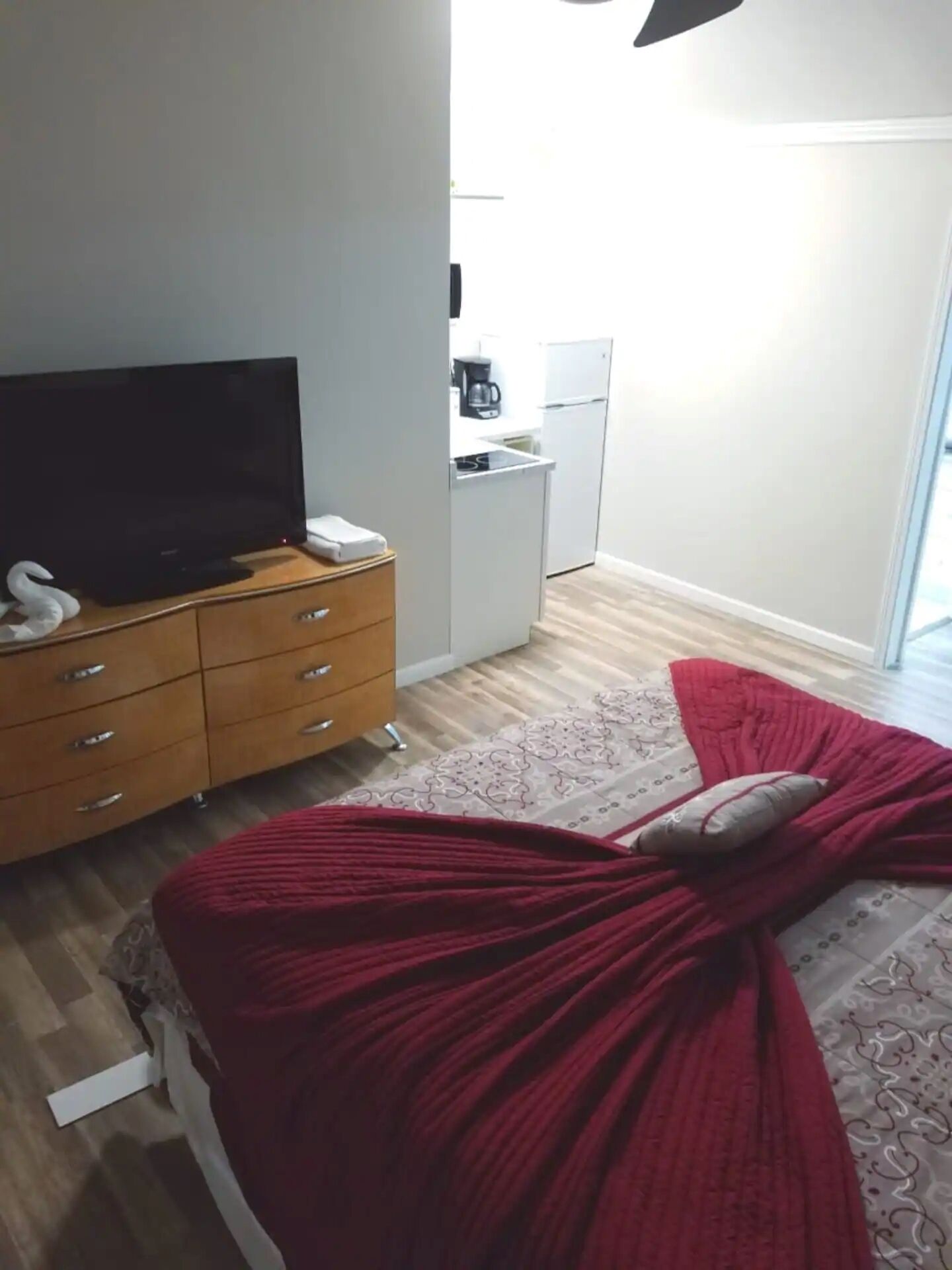 Ein Schlafzimmer mit einem Bett, einem Fernseher auf einer Holzkommode, einer Küchenzeile mit Kühlschrank und Kaffeemaschine sowie einem gemusterten Teppich.