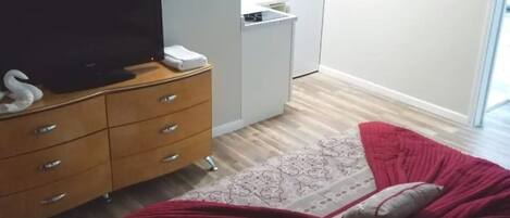 Ein Schlafzimmer mit einem Bett, einem Fernseher auf einer Holzkommode, einer Küchenzeile mit Kühlschrank und Kaffeemaschine sowie einem gemusterten Teppich.