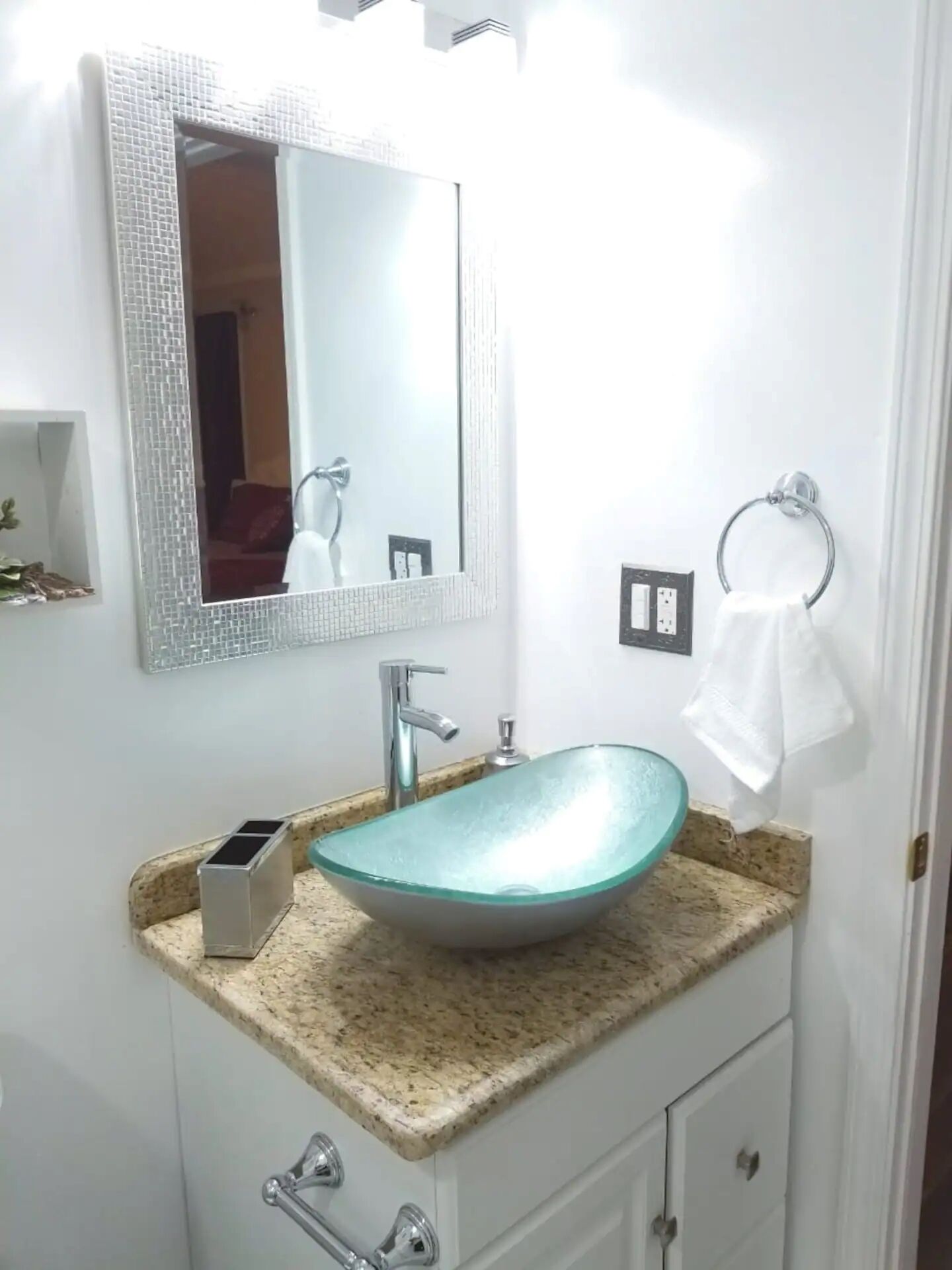 A bathroom with a glass sink, a mirror, a towel ring, and a small shelf.