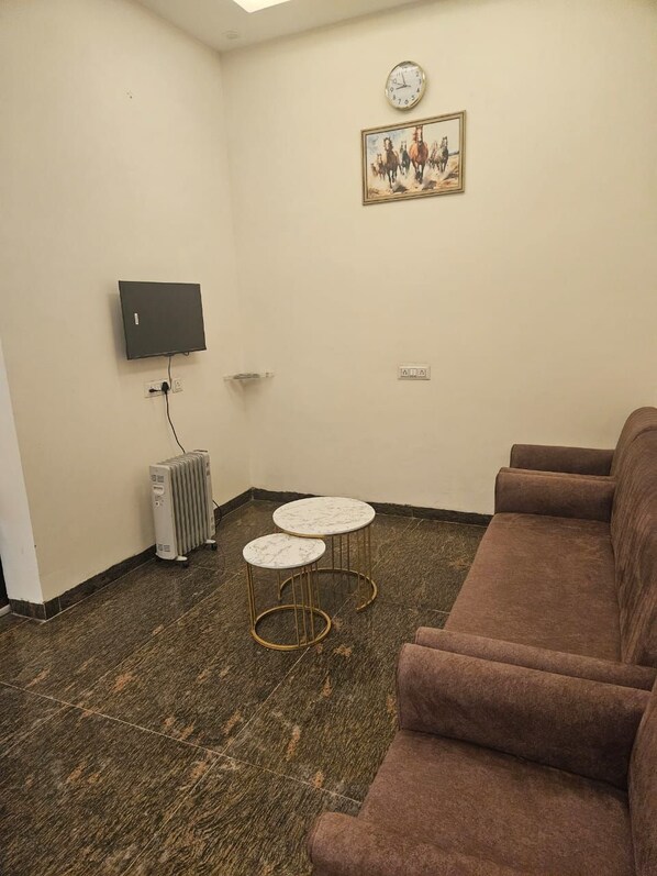 Une pièce avec un canapé marron, deux tables surmontées de marbre, un radiateur, un téléviseur accroché au mur et un tableau représentant des chevaux.