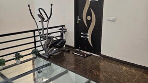Een kamer met een glazen tafel, fitnessapparatuur, een deur met decoratieve details en een klein fotolijstje aan de muur.