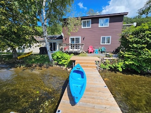 Un muelle de madera que lleva a una casa con antena parabólica, un kayak y mobiliario exterior.