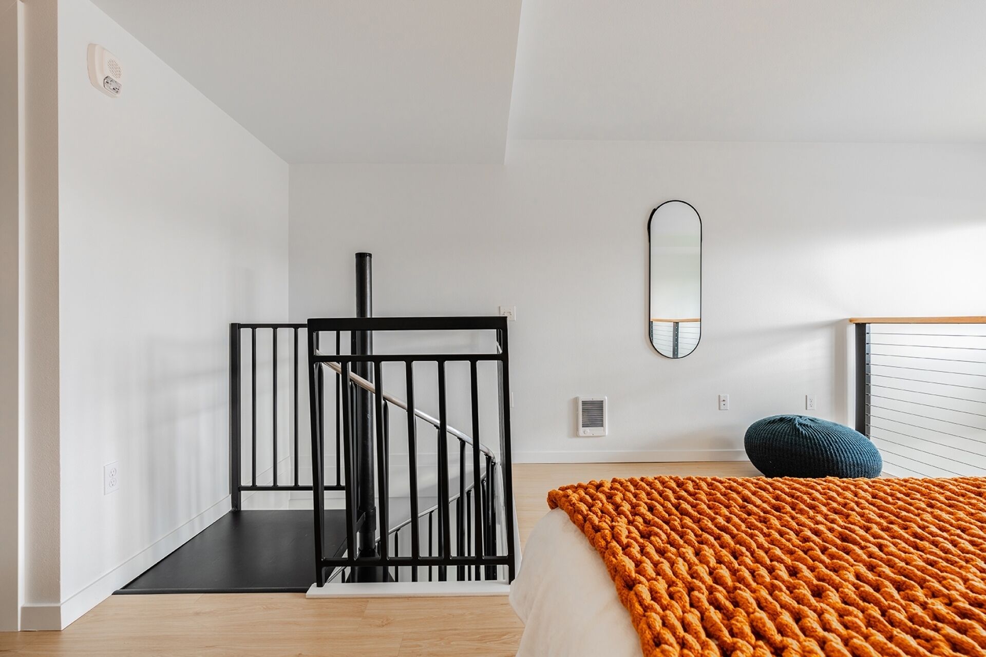 A modern bedroom with a staircase, a bed with an orange blanket, a mirror, and a window with blinds.
