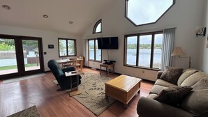 A spacious living room with a large octagonal window, a flat-screen TV, a wooden coffee table, and a sofa. - UP Lake Home with access to 3 lakes - fish, swim, relax! WIFI (Vulcan)