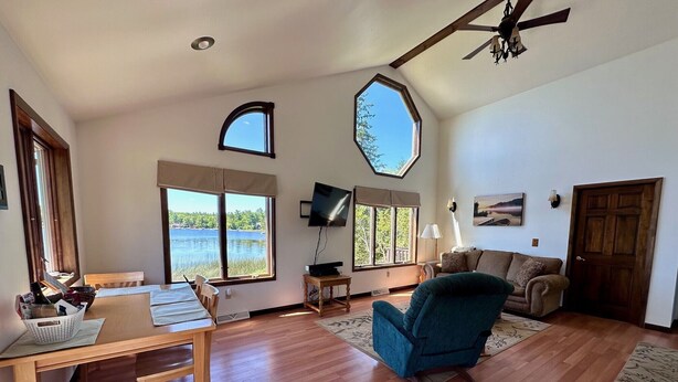 A spacious living room with a wooden floor, a blue armchair, a wooden dining table, and a TV. There are large windows offering a view of a lake and trees. - UP Lake Home with access to 3 lakes - fish, swim, relax! WIFI (Vulcan)