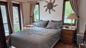 A bedroom with a large bed, a nightstand with a lamp, a wooden chair, and a view of trees through the windows. - UP Lake Home with access to 3 lakes - fish, swim, relax! WIFI (Vulcan)