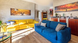 Un salon avec un canapé jaune, un canapé bleu, une table basse en verre et une bibliothèque.