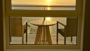 Vista desde una ventana que da a una terraza de madera con una mesa redonda y dos sillas, con vistas a un cuerpo de agua al atardecer.