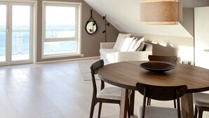 Un loft espacioso con techo alto, comedor con mesa y sillas de madera, sofá blanco y ventana grande con vista a un cuerpo de agua.