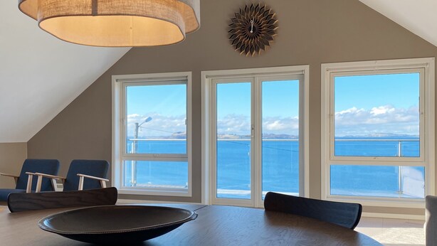 Un comedor con una mesa redonda, sillas y una lámpara colgante. Se ven amplias ventanas con vistas al océano y a las montañas.