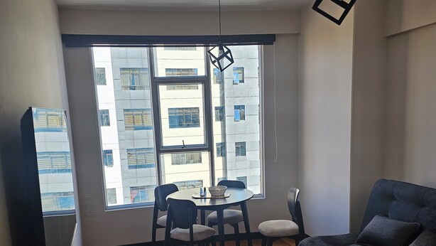 Una sala de estar moderna con un sofá, una mesa de comedor con sillas, un televisor y un ventanal con vistas a edificios.