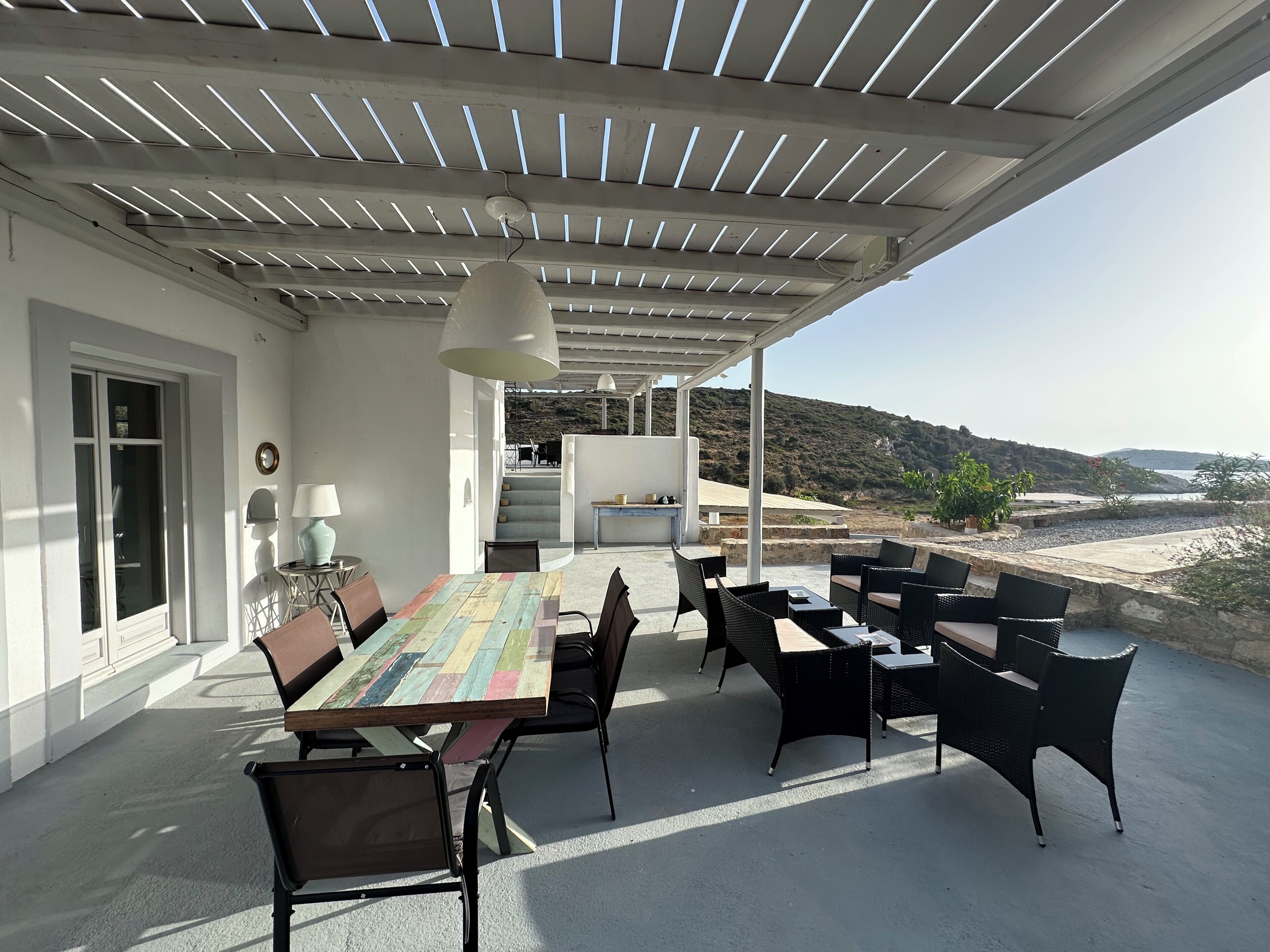 Une terrasse extérieure avec une table et des chaises, un espace couvert offrant une vue sur la mer, et un escalier menant à un niveau supérieur.