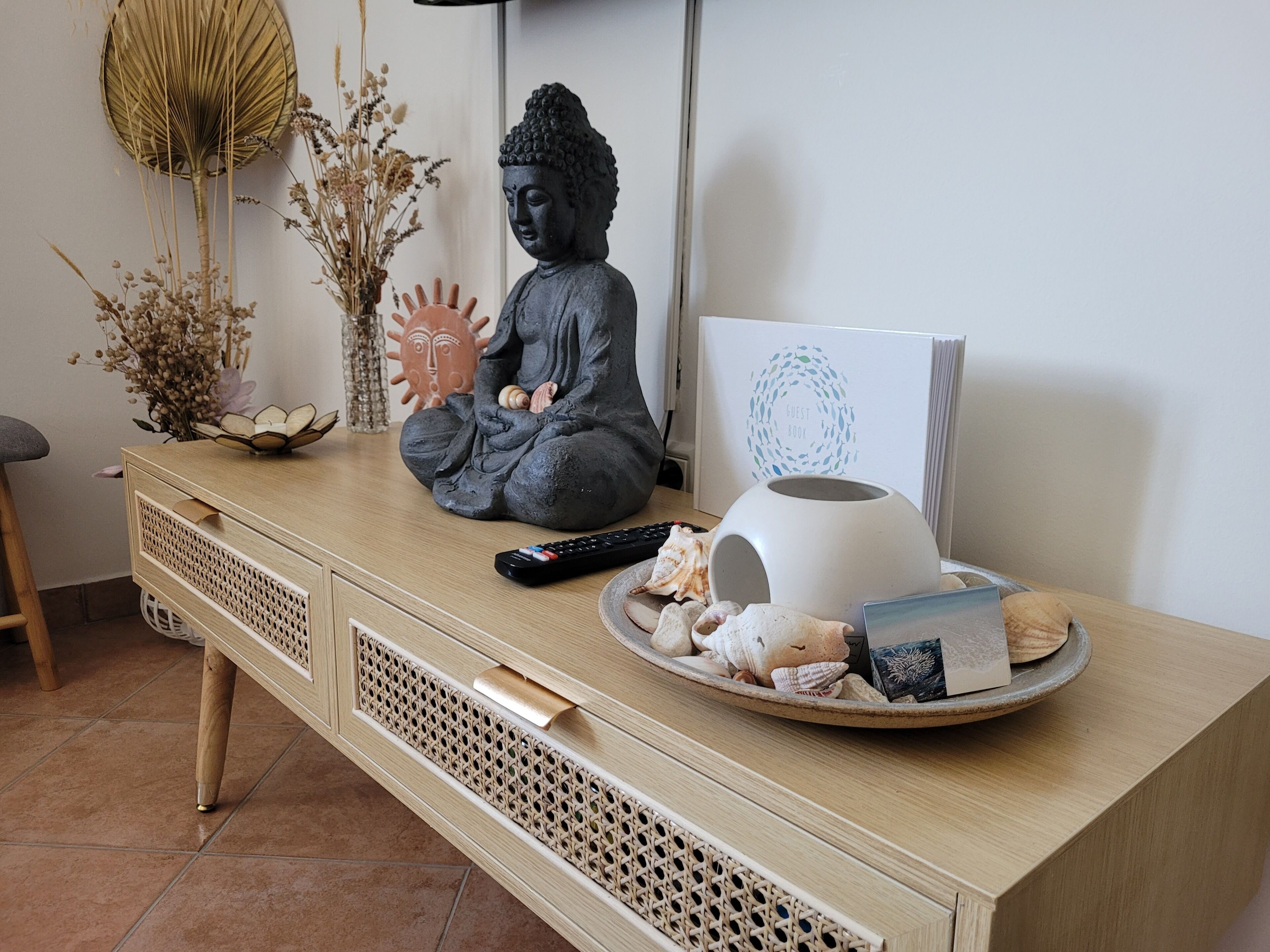 A wooden desk with a Buddha statue, a vase with dried flowers, a remote control, and a tray with seashells and a book.