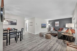 A modern living room with a dining area, featuring a grey wooden floor, contemporary furniture, and artwork on the walls.