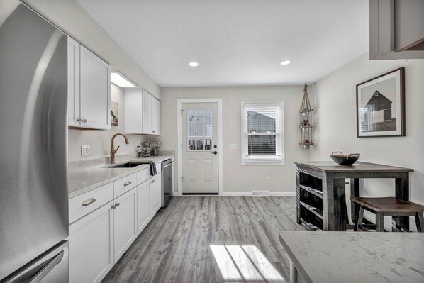 A kitchen with white cabinets, a stainless steel refrigerator, a wooden dining table, and a window with blinds.