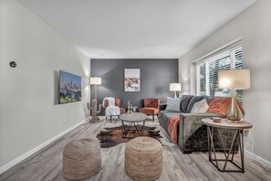 A modern living room with a cityscape painting, a large window with blinds, a grey sofa, a round coffee table, and two lamps.