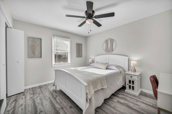 A bedroom with a bed, a ceiling fan, a window with blinds, a bedside table with a lamp, and a chair.