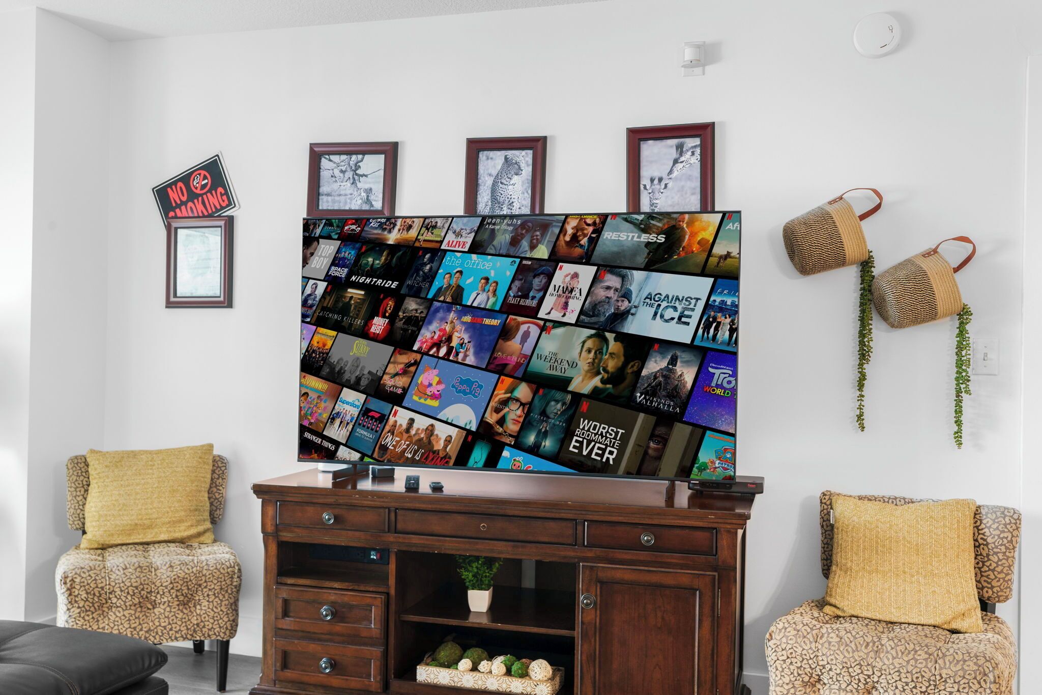 A living room with a flat-screen TV displaying movie posters, a wooden cabinet, two patterned armchairs, and a 'No Smoking' sign.