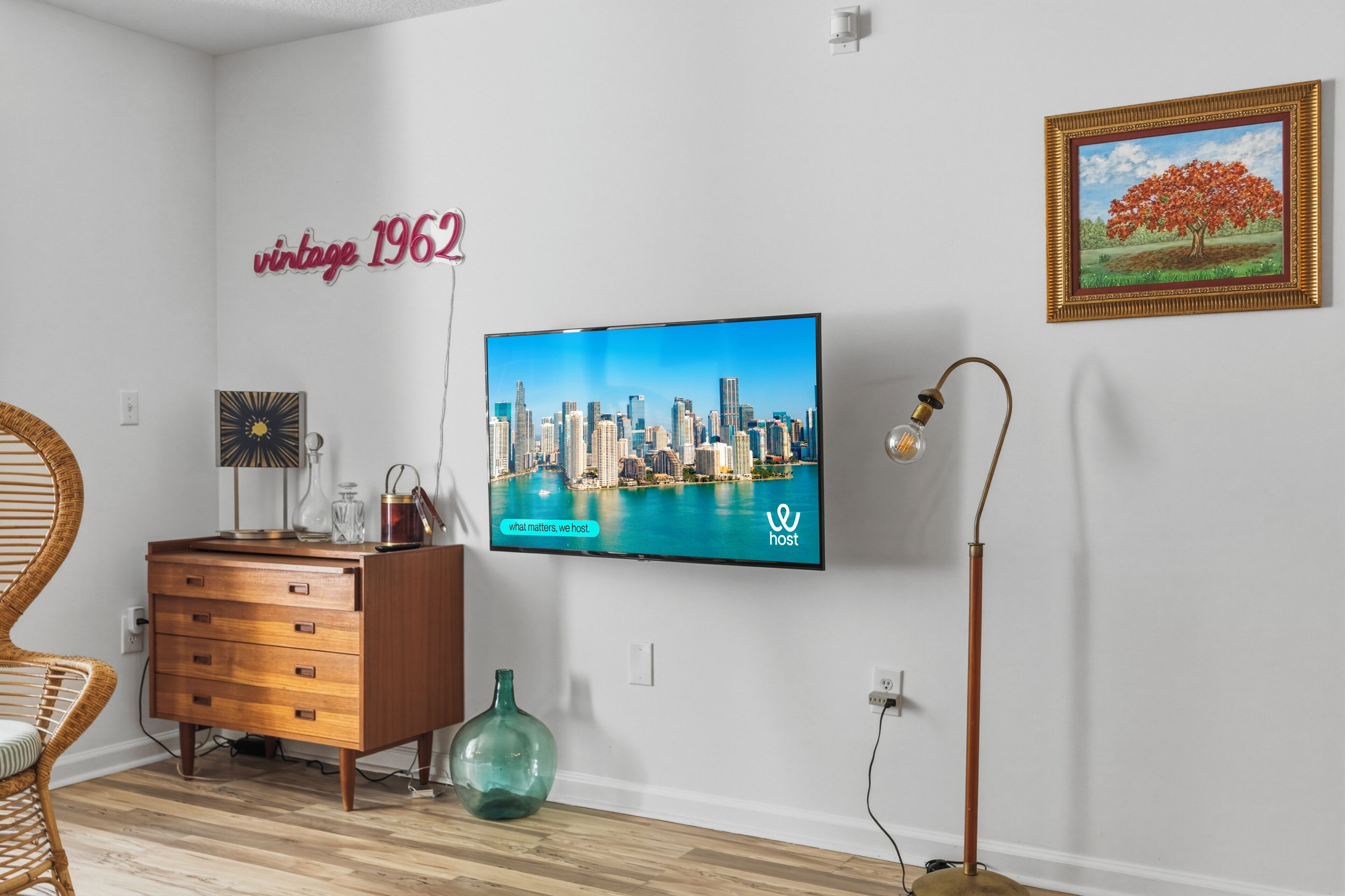 A modern living room with a flat-screen TV, vintage 1962 wall art, a framed painting, a wooden dresser, and a floor lamp.