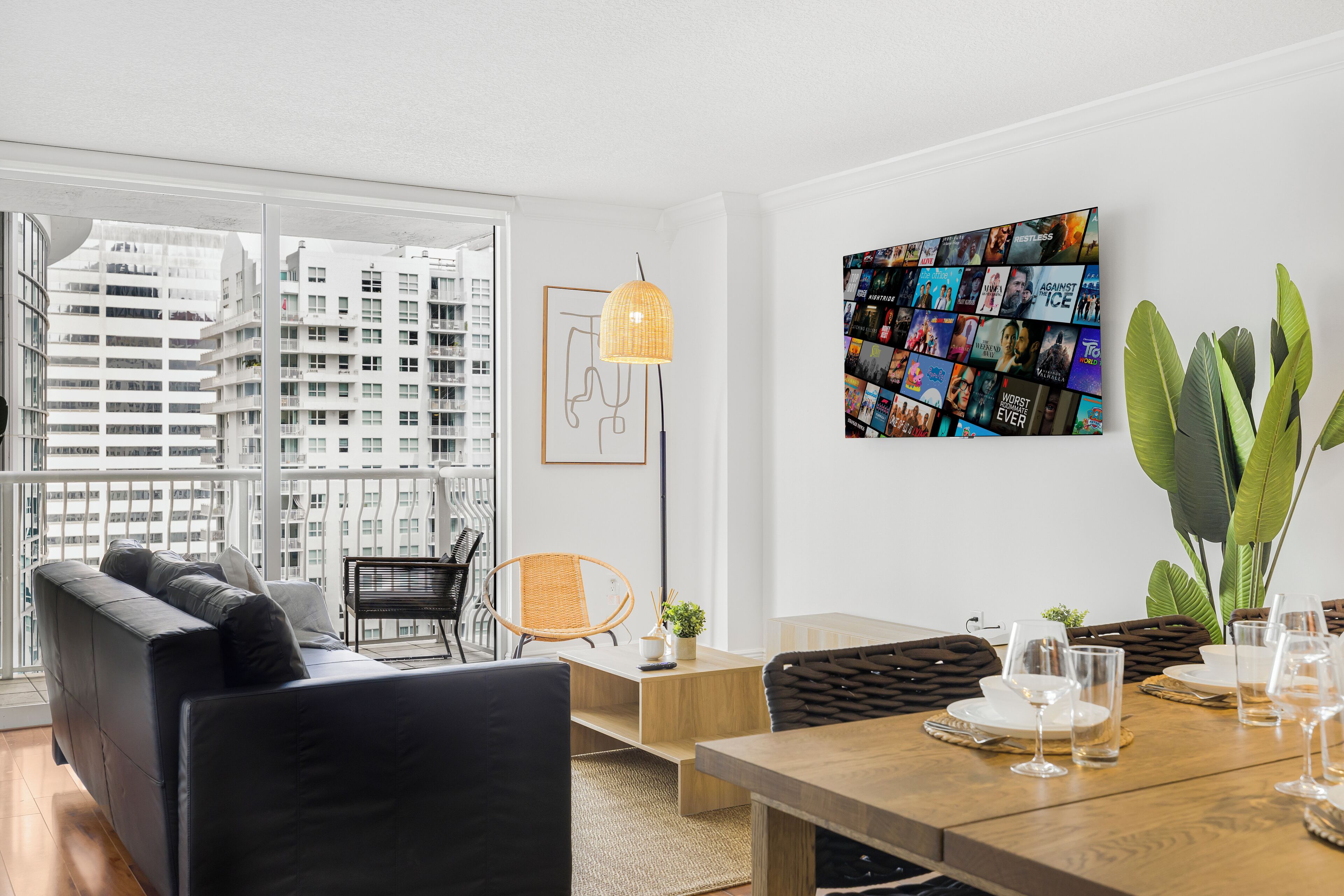 Ein modernes Wohnzimmer mit einem großen Fenster, einem schwarzen Ledersofa, einem Esstisch gedeckt mit Gläsern und einer Wandcollage.