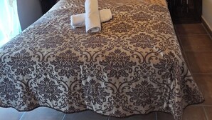 Una cama bien tendida con una colcha estampada, una toalla enrollada y una sola almohada. Hay dos mesitas de noche, una con una lámpara y la otra con un enchufe.
