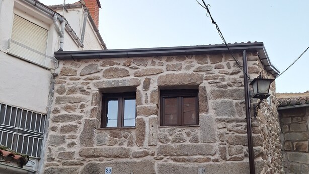 Un edificio de piedra con una puerta marrón y dos ventanas, un pequeño balcón y una caja de servicios en la planta baja.