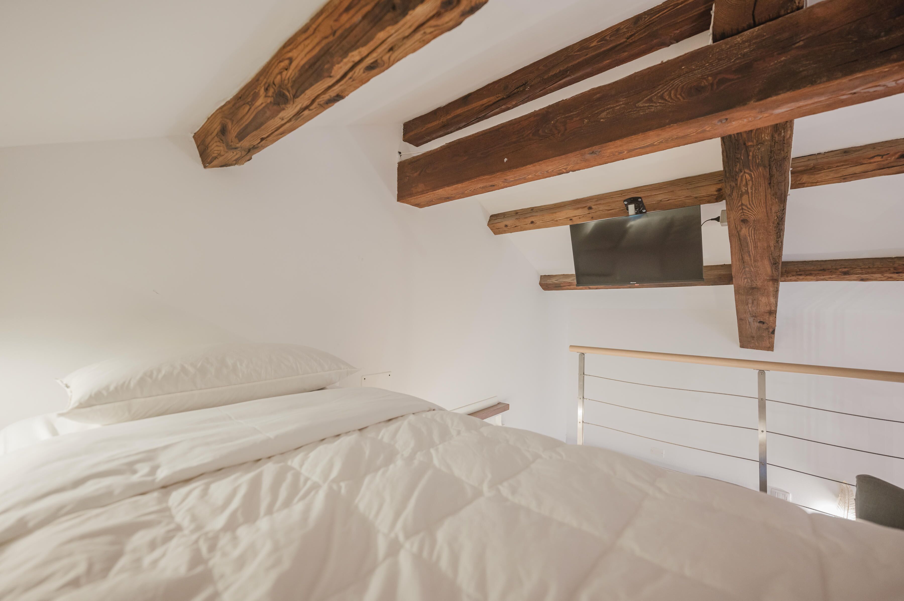 A bedroom with a bed, wooden beams, a metal headboard, and a small shelf.