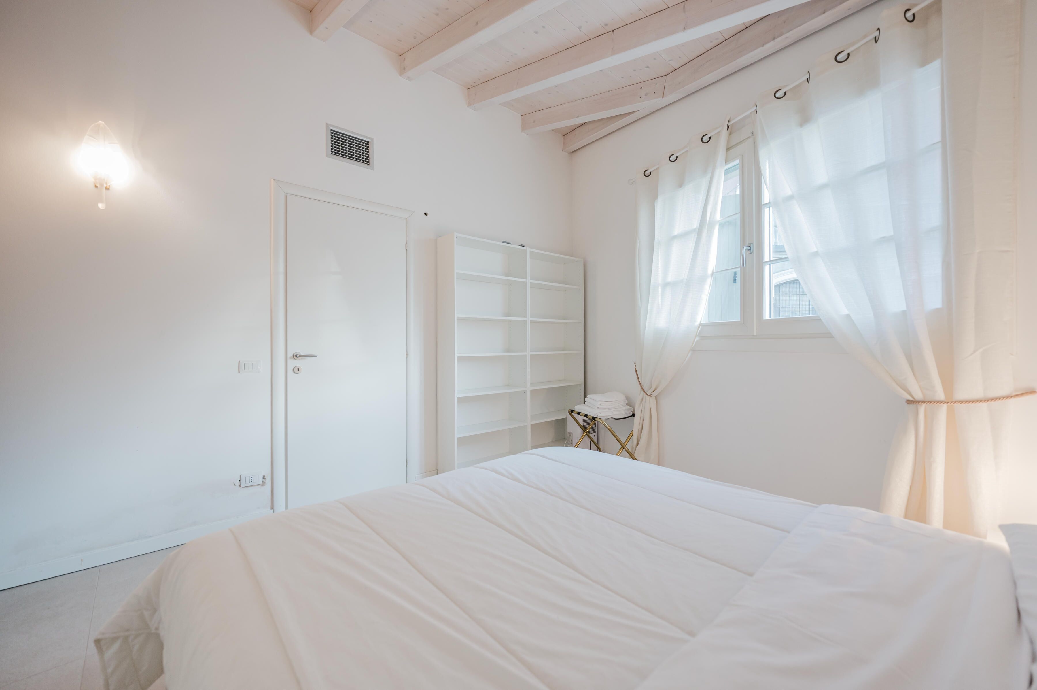 Une chambre avec un grand lit, une armoire, une petite table avec une serviette et une fenêtre ornée de rideaux légers.
